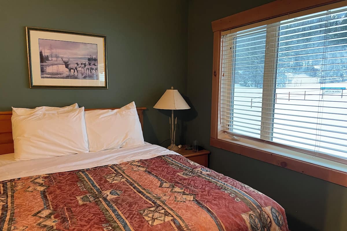 A bed with white linens, a patterned red and beige bedspread, a framed picture on the wall, a bedside lamp, and a window with blinds.