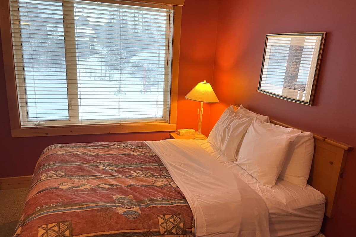 A hotel room with a bed, a nightstand, a lamp, and a window with blinds.