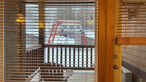 A wooden-framed window with horizontal blinds, a metal grill, and a view of a red ladder outside.