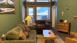 A living room with a green couch, a brown armchair, a wooden coffee table, and a lamp. There is a painting of a mountain on the wall and a wooden cabinet with baskets.