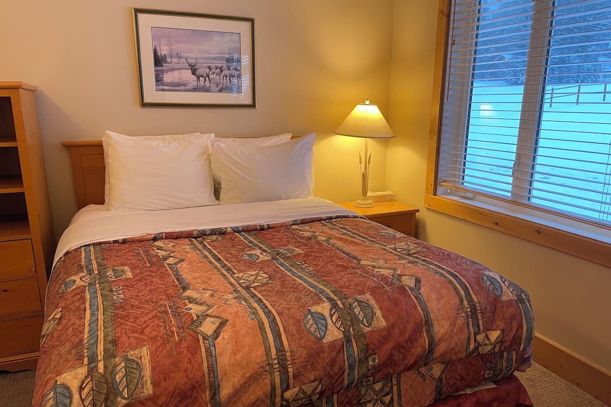 A neatly made bed with a patterned bedspread, a framed picture on the wall, a bedside lamp, and a window with blinds.