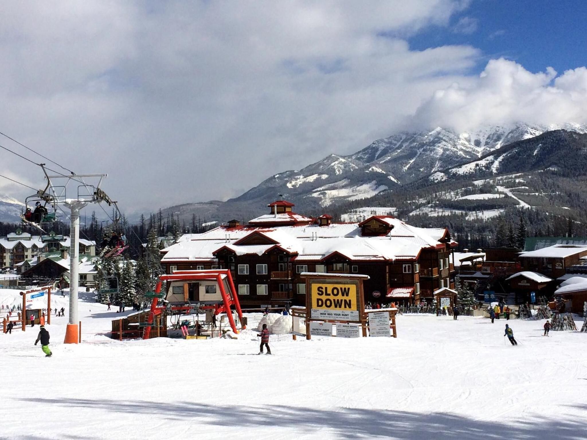 Ein Skigebiet mit einem 'SLOW DOWN'-Schild, Skipisten und schneebedeckten Bergen im Hintergrund.