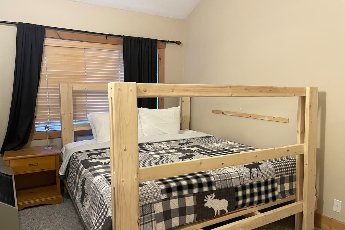 A single bed with a wooden frame, a bedside table, a lamp, and a window with blinds.
