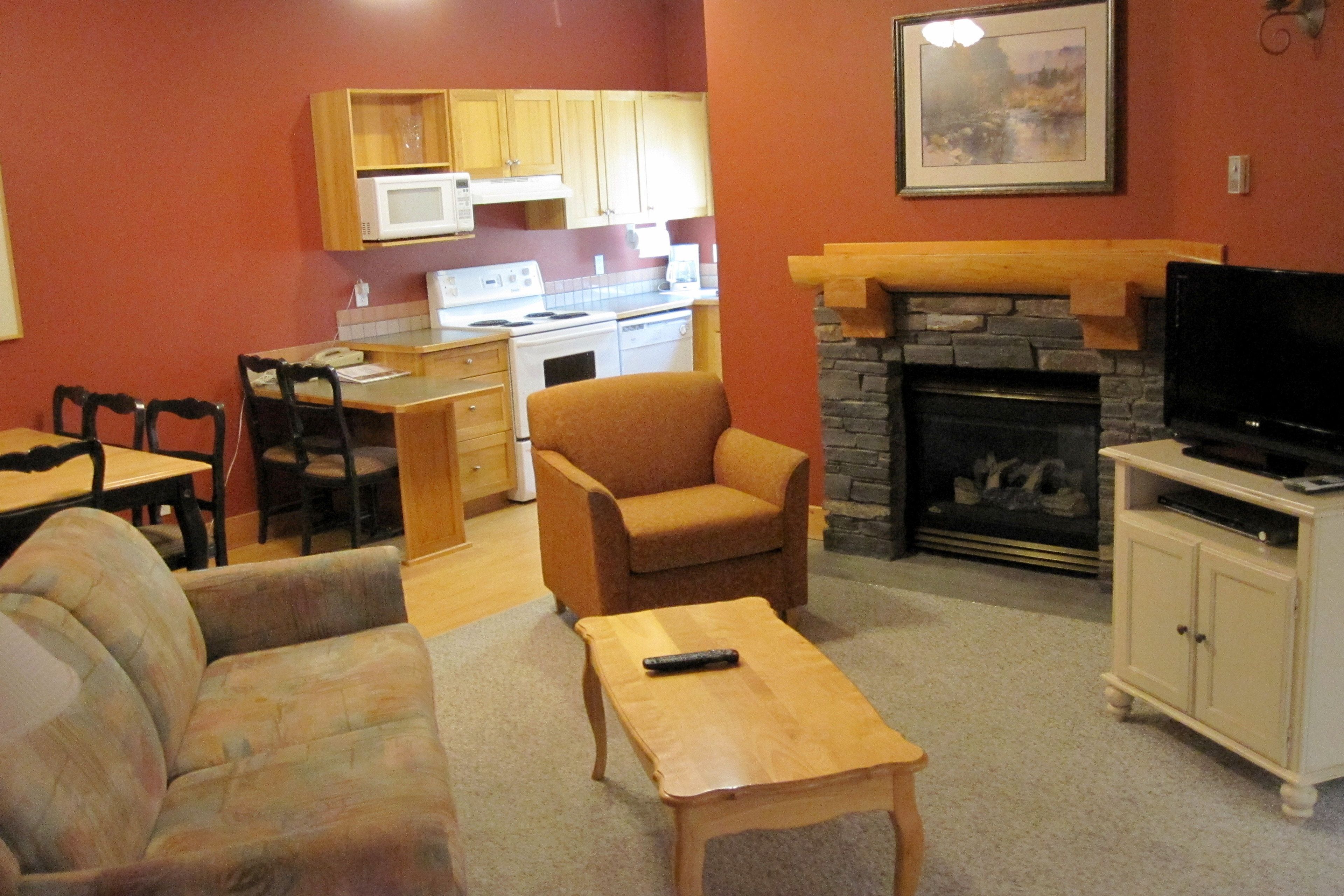 A living room with a sofa, armchair, coffee table, and a fireplace. There is a kitchen area with wooden cabinets and a microwave.