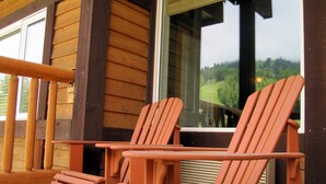 Two wooden Adirondack chairs and a table outside a cabin with a large window overlooking a forested area.