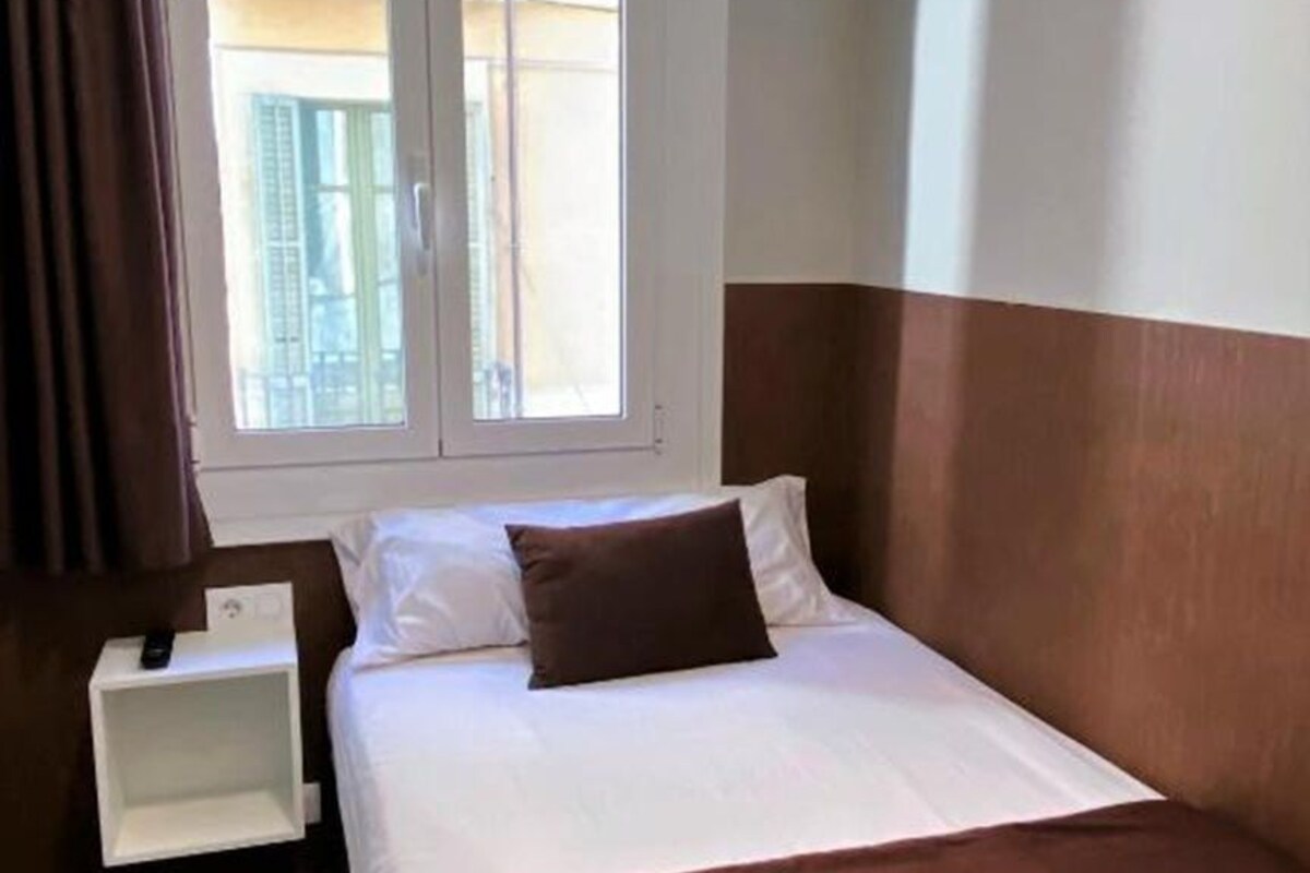 Una habitación de hotel con cama, mesita de noche, ventana con vistas a edificios y cortina.