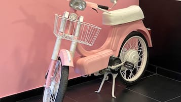 Una patineta rosa con cesto y asiento blanco, estacionada en el interior frente a una pared rosa.