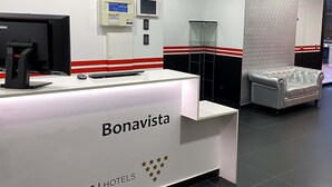 Een receptiebalie met de tekst 'Bonavista BCN URBANNESS | HOTELS', een computermonitor, een kleine koelkast en een glazen scheidingswand.