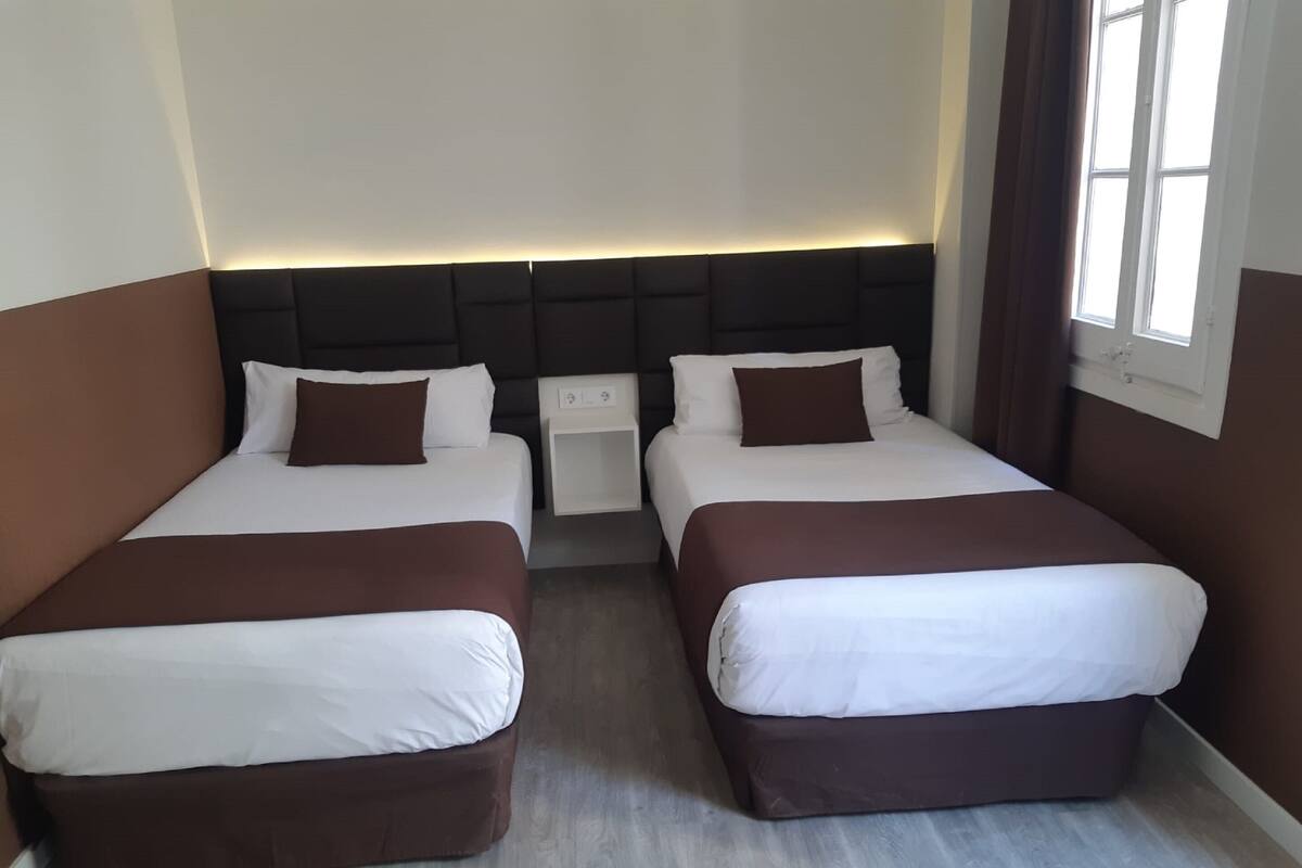 A hotel room with two beds, each with a brown headboard, white bedding, and a bedside table with a lamp.