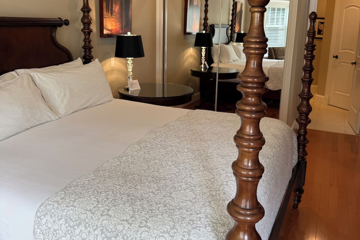 A neatly made bed with a patterned bedspread, a wooden four-poster headboard, a bedside table with a lamp, and a mirror reflecting the room.