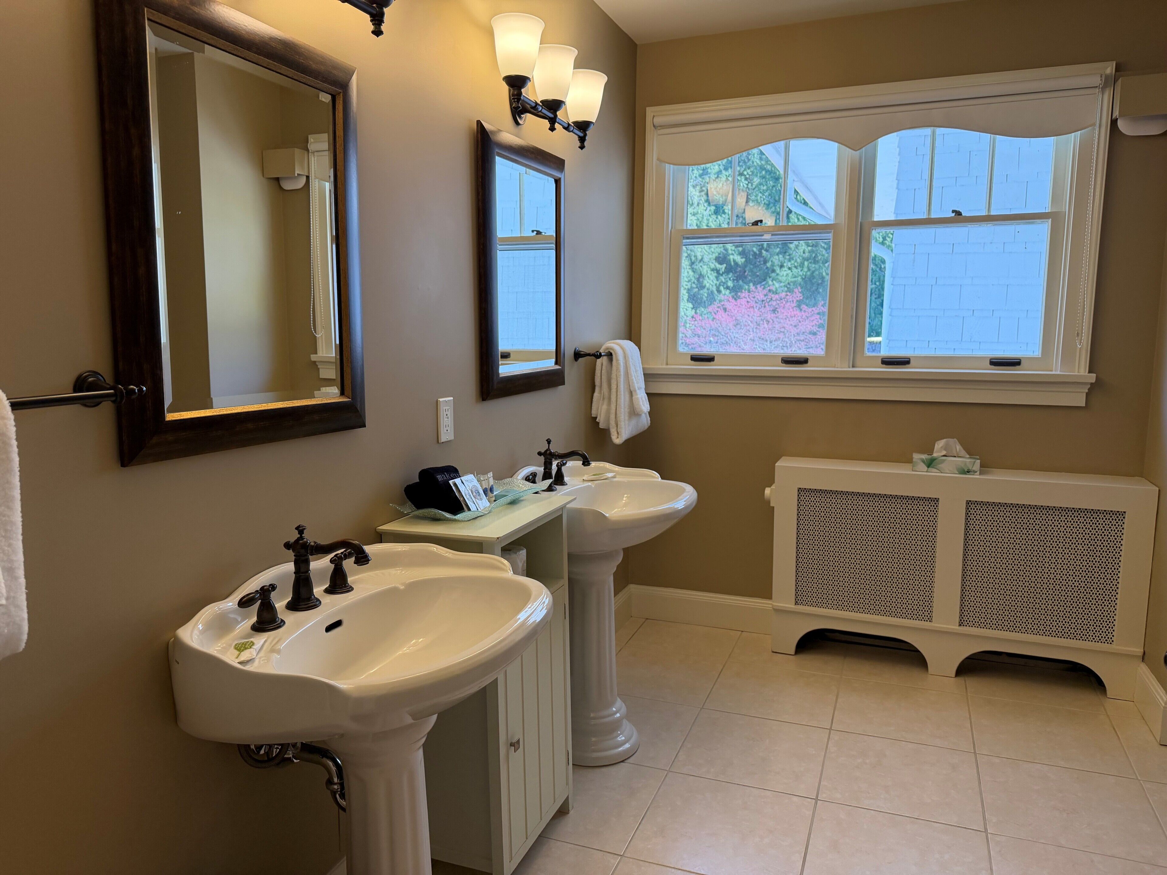 A bathroom with two sinks, a mirror, wall sconces, and a window.