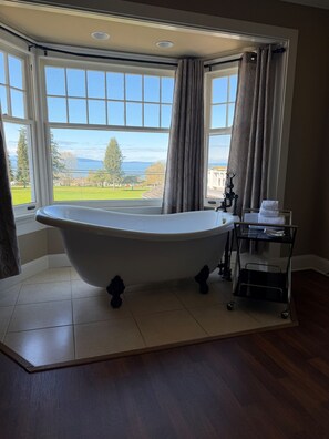 A corner bathroom with a freestanding bathtub, a vanity with a towel rack, and a view of a green landscape through large windows.