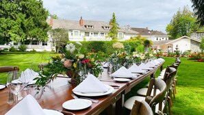 A long dining table set outdoors with chairs, surrounded by lush greenery and a white building in the background. - Crown Mansion Boutique Hotel & Villas (Qualicum Beach)