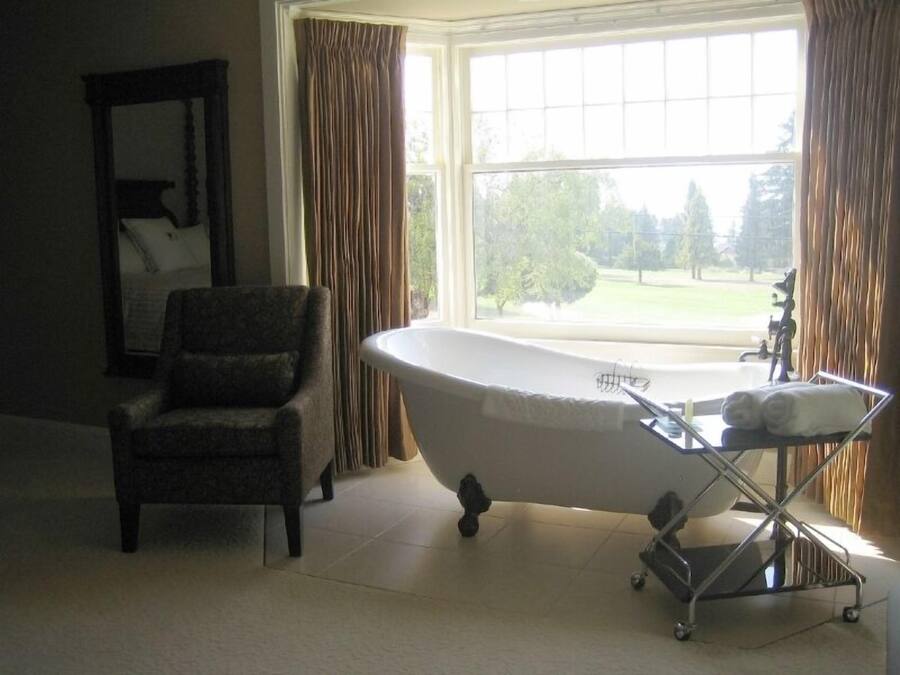 Un baño con una bañera independiente, una silla y un ventanal con cortinas.