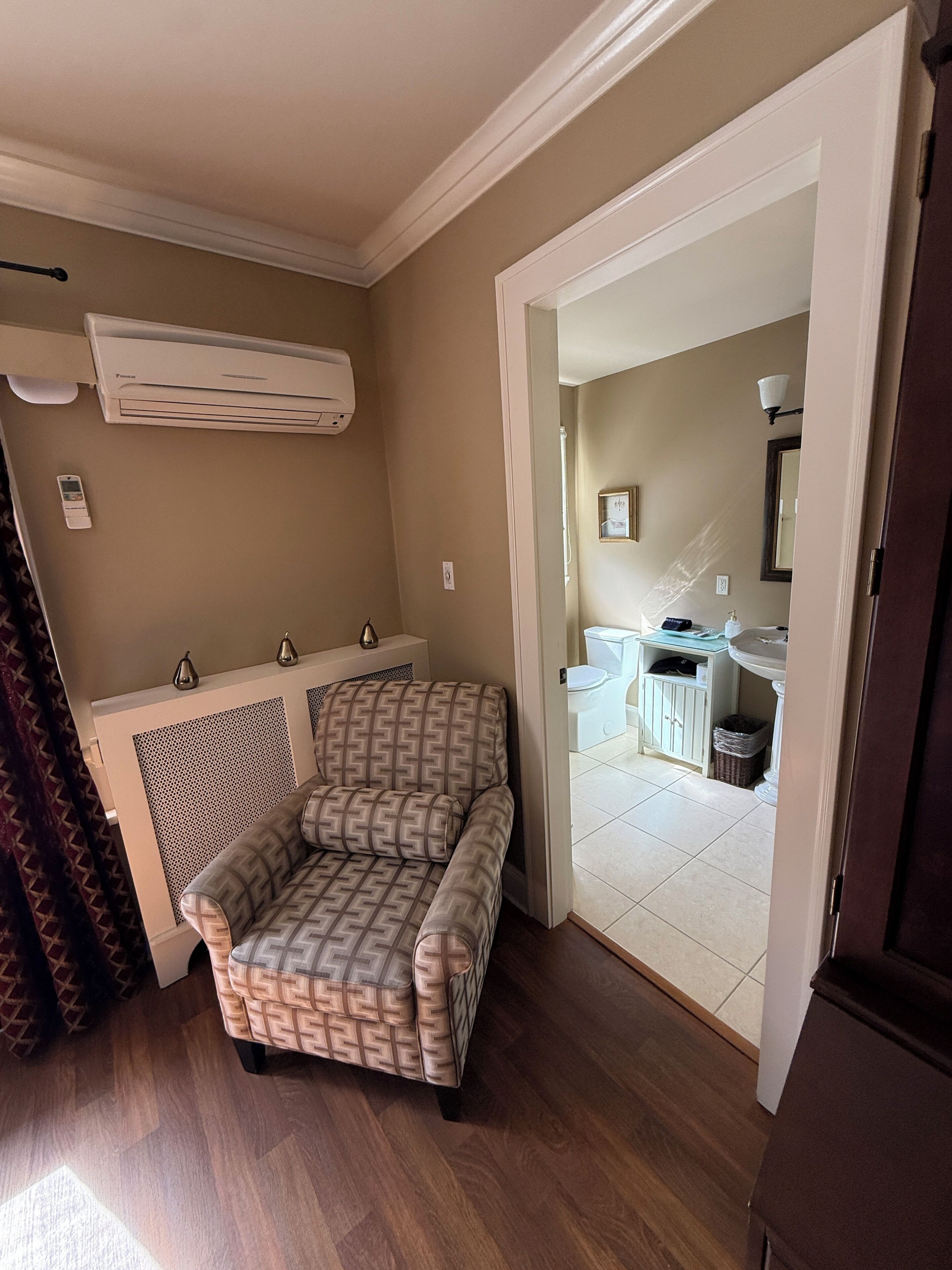 A room with a patterned sofa, a white radiator, a bathroom visible through an open door, and a tiled floor.