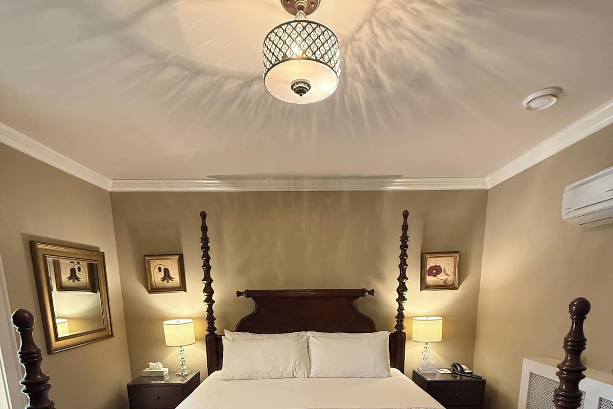A traditional four-poster bed with a floral bedspread, two bedside lamps, a ceiling light fixture, and a wooden bench at the foot of the bed.