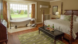 A bedroom with a large window, a bathtub, a bed with a wooden headboard, a bedside table with a lamp, and a green rug. - Crown Mansion Boutique Hotel & Villas (Qualicum Beach)