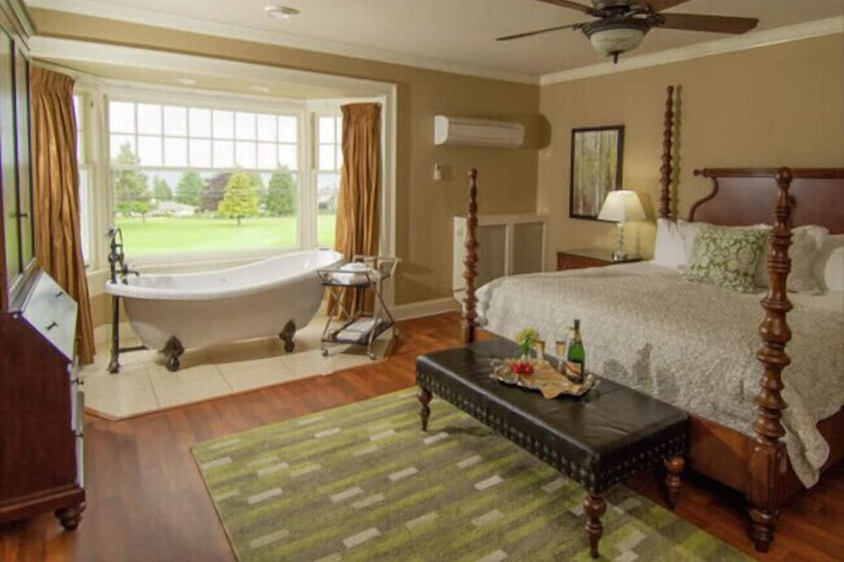 A bedroom with a large window, a bathtub, a bed with a wooden headboard, a bedside table with a lamp, and a green rug.