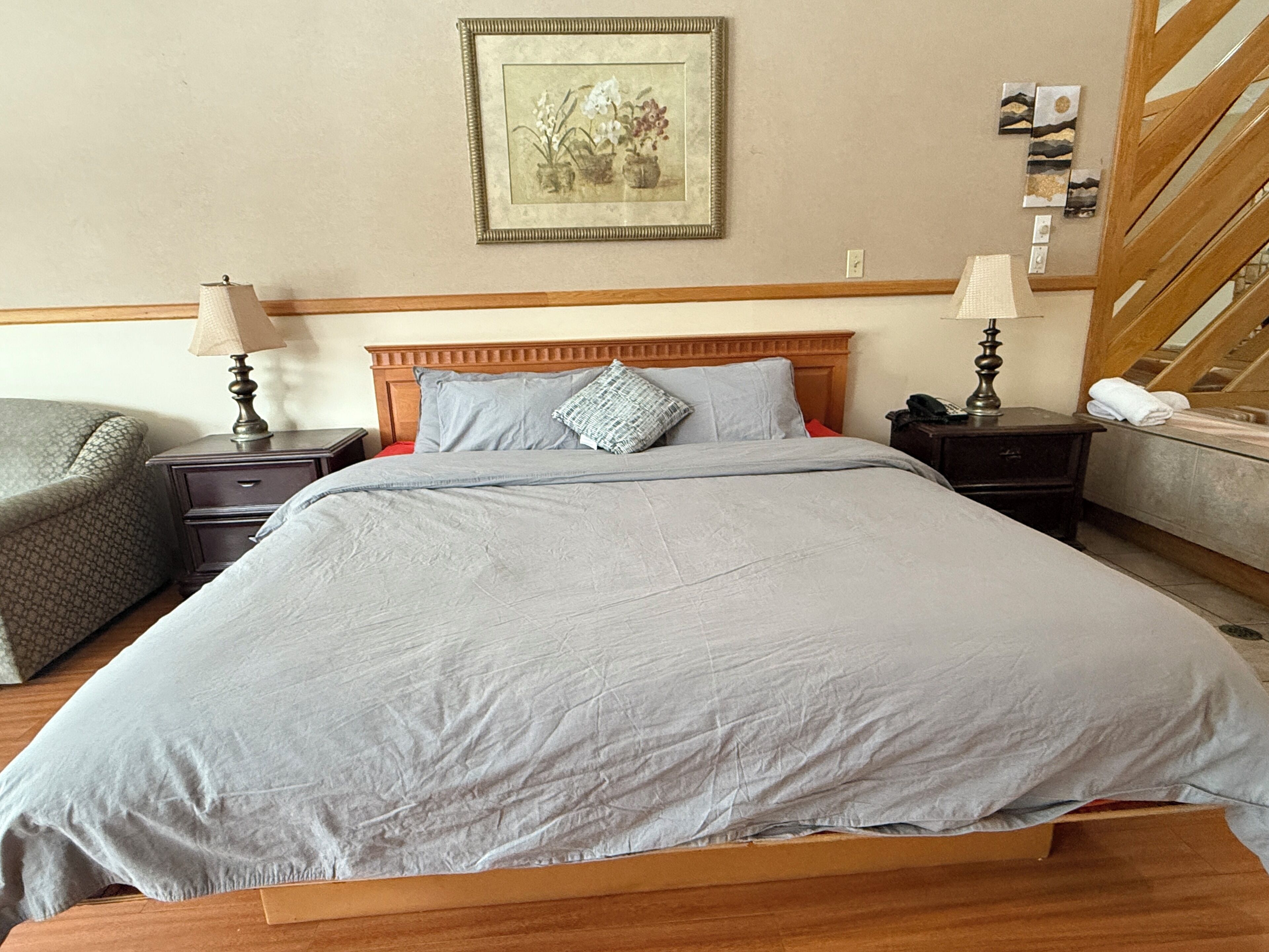 A neatly made bed with a wooden headboard, two bedside tables with lamps, a framed artwork on the wall, and a staircase to the right.