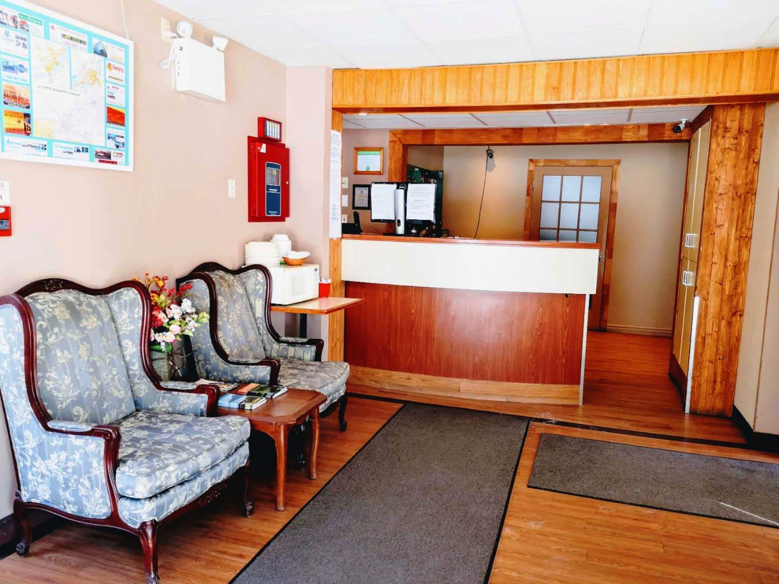 Un espace d’accueil avec un comptoir en bois, deux fauteuils rembourrés, une petite table ornée d’un vase de fleurs et un tableau d’affichage fixé au mur.