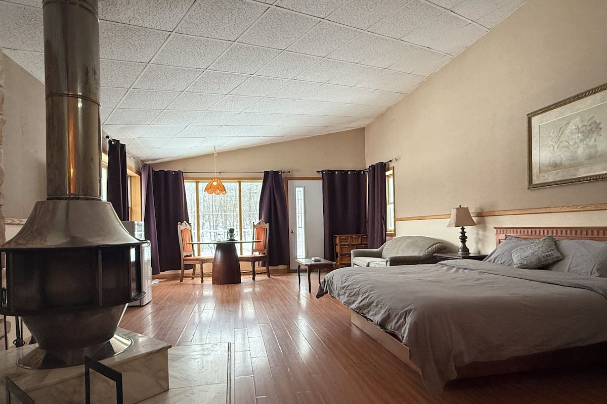 Hotellihuone, jossa on suuri sänky, puulattia, takka, ruokapöytä tuoleineen ja ikkuna, jossa on verhot.