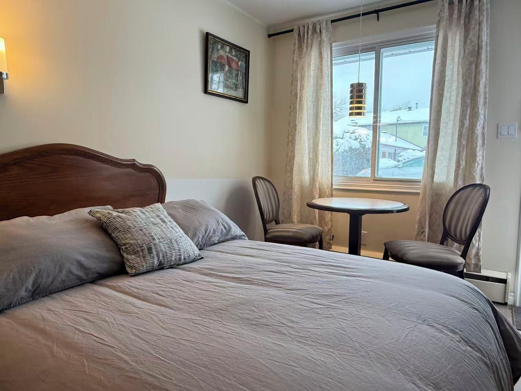 A hotel room with a large bed, two chairs, a small round table, a window with curtains, and a wall-mounted picture.