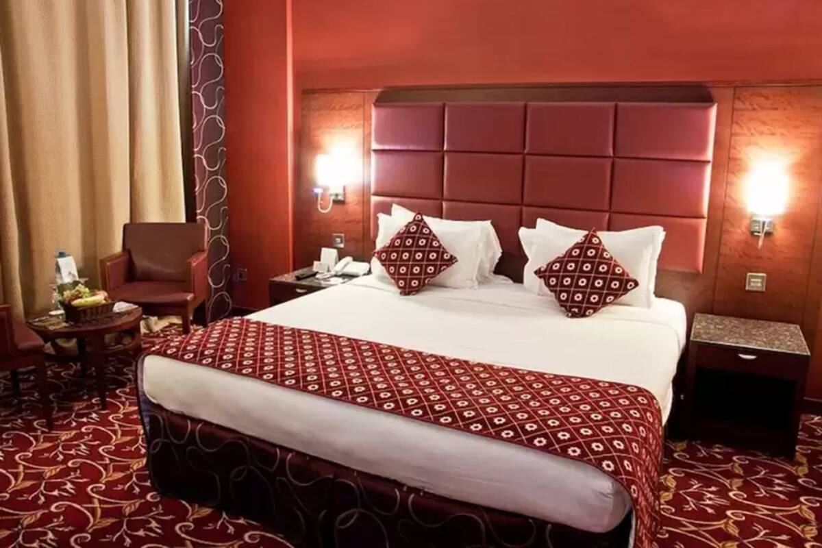 A hotel room with a large bed, a red patterned bedspread, a red upholstered bench, a wooden headboard, two bedside lamps, a nightstand, a chair, and a small table with a lamp.