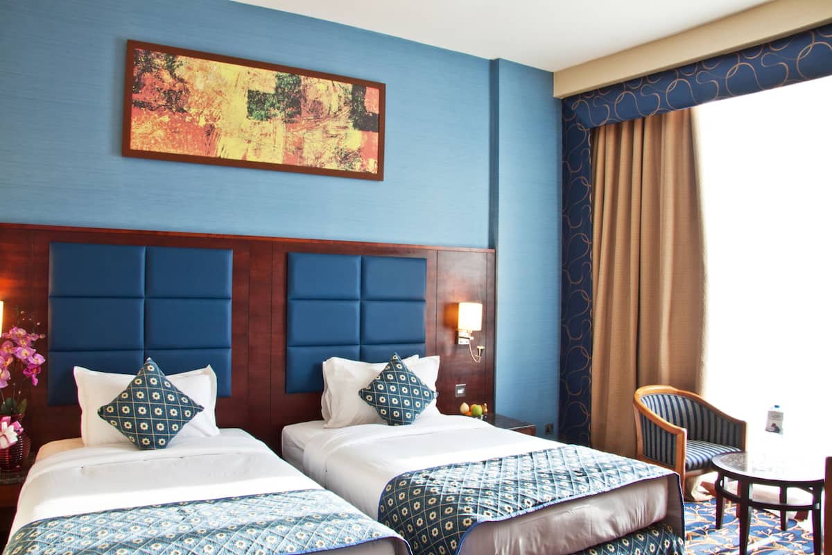 A hotel room with two beds, a headboard, a wall-mounted painting, a window with curtains, a chair, and a small table.