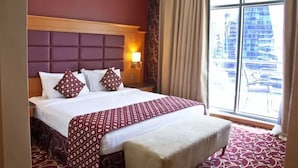 A hotel room with a large bed, a headboard, a bedside table with a lamp, a window with curtains, and a view of buildings outside. - Ramee Rose Hotel (Dubai)
