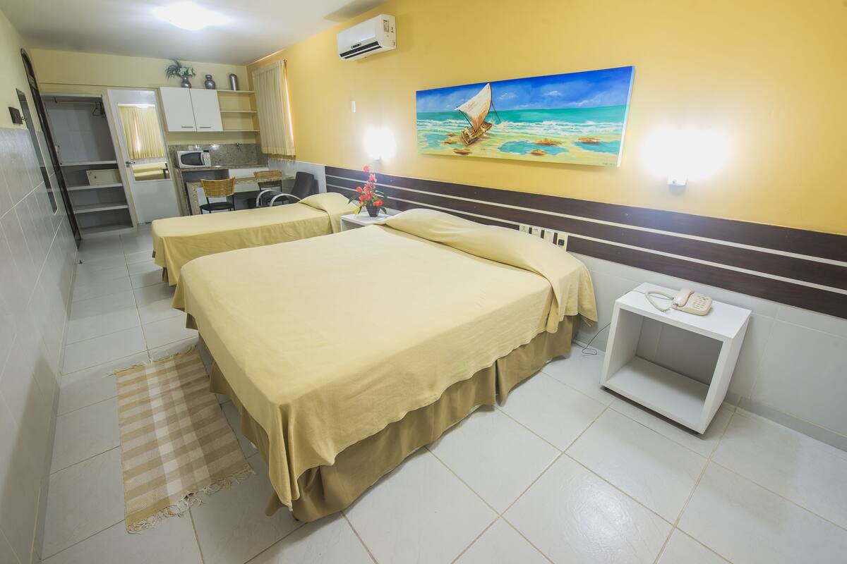 Une chambre d’hôtel avec deux lits, un coin cuisine, un tableau représentant un voilier et une petite table avec un téléphone.
