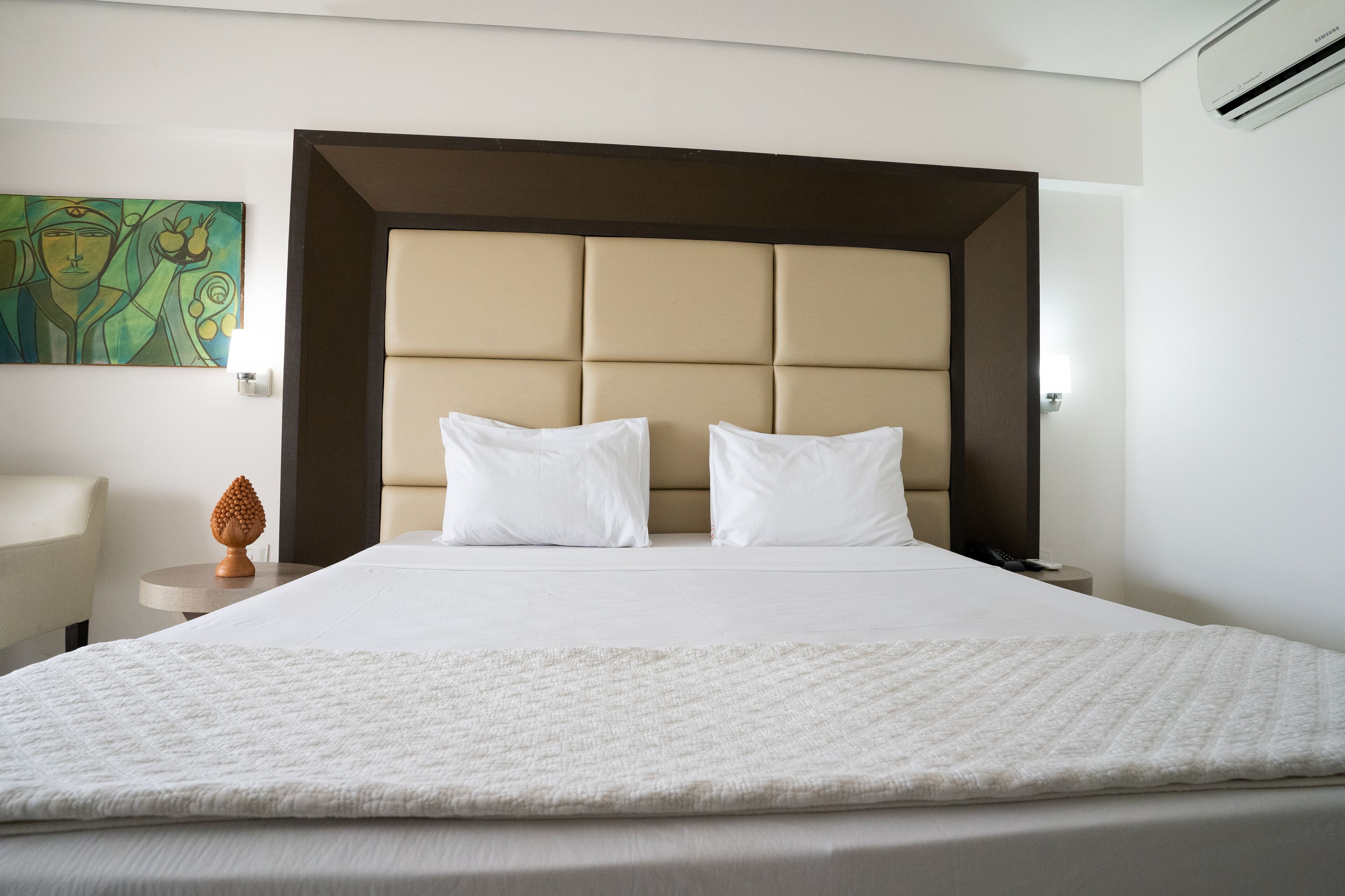 A hotel room with a large bed, a headboard, a bedside table, a lamp, and a wall art piece.