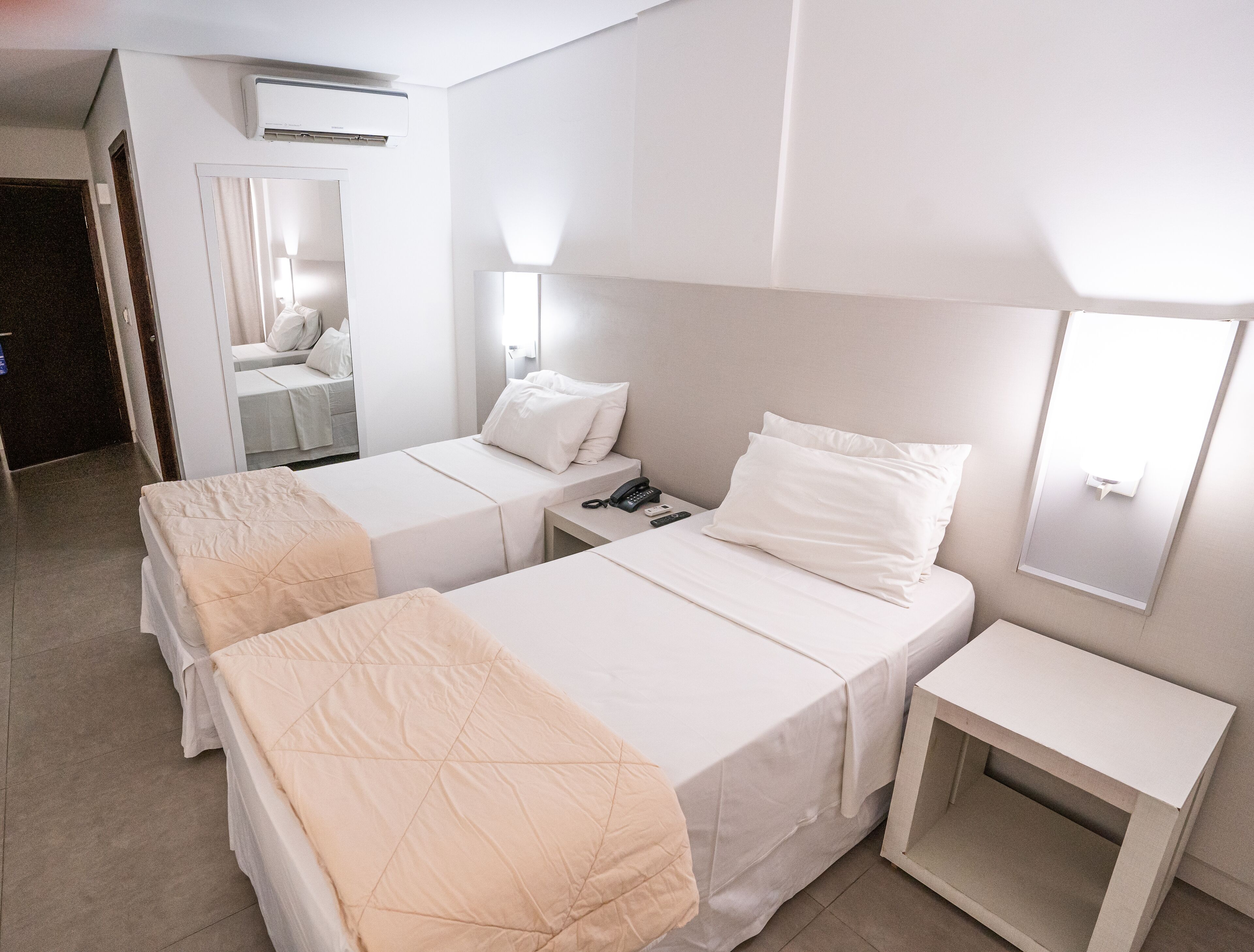 A hotel room with two beds, a bedside table, a nightstand, a mirror, and an air conditioning unit.