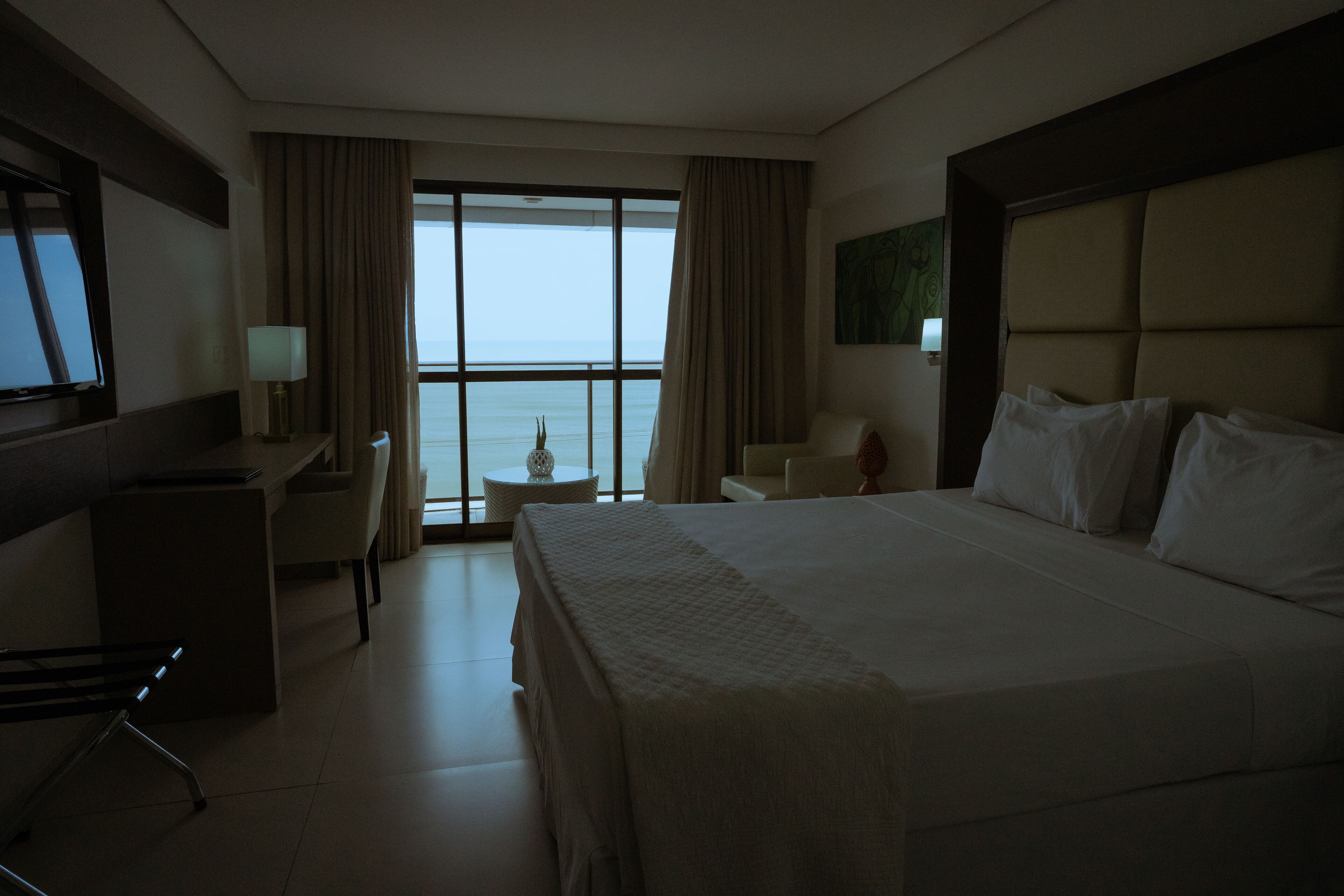 A hotel room with a large bed, a desk, a chair, a small table, and a view of the sea through the window.