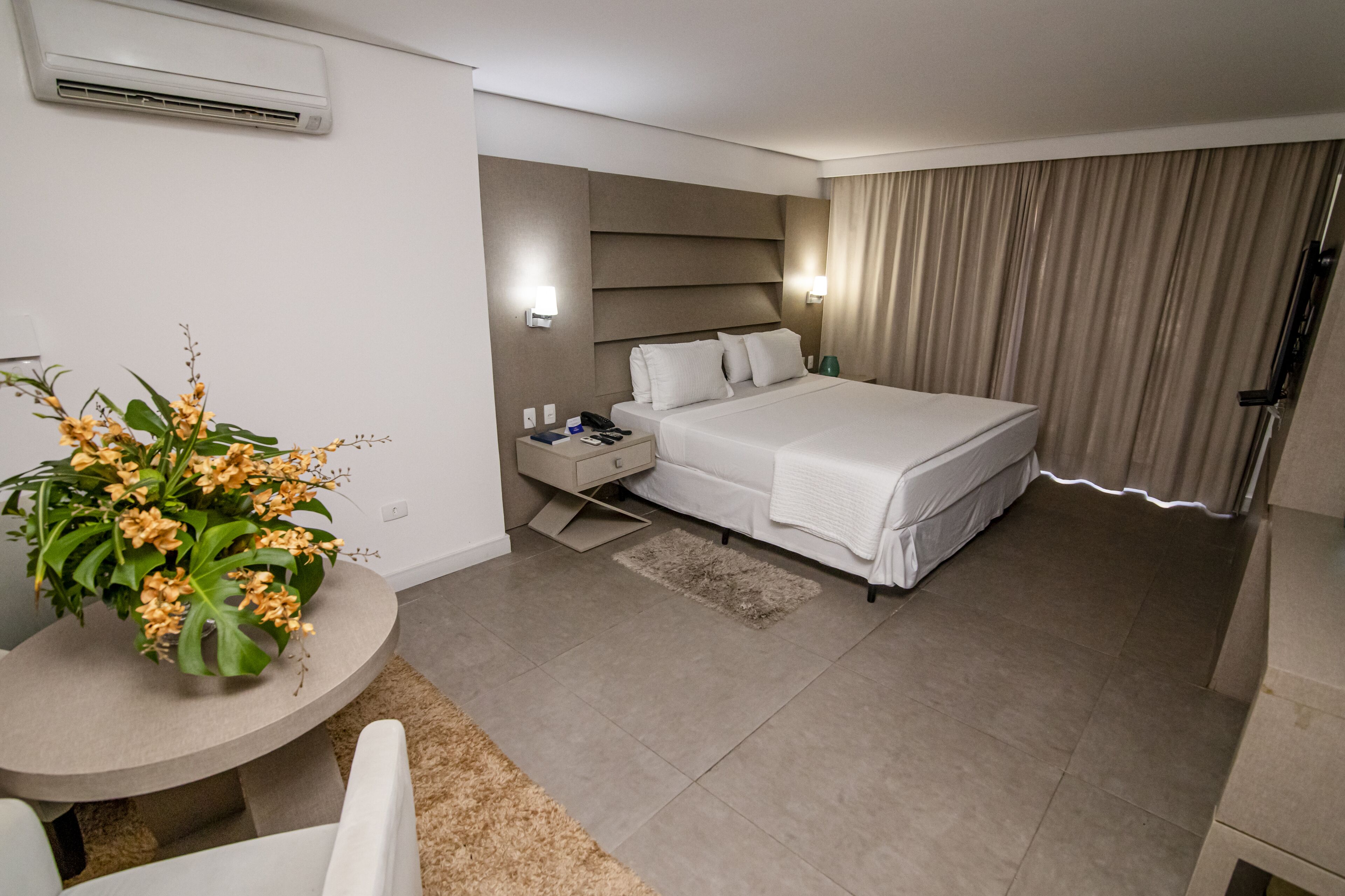 Ein modernes Hotelzimmer mit einem großen Bett, Nachttischen, einem kleinen runden Tisch mit einer Pflanze und Blick auf die Stadt durch das Fenster.