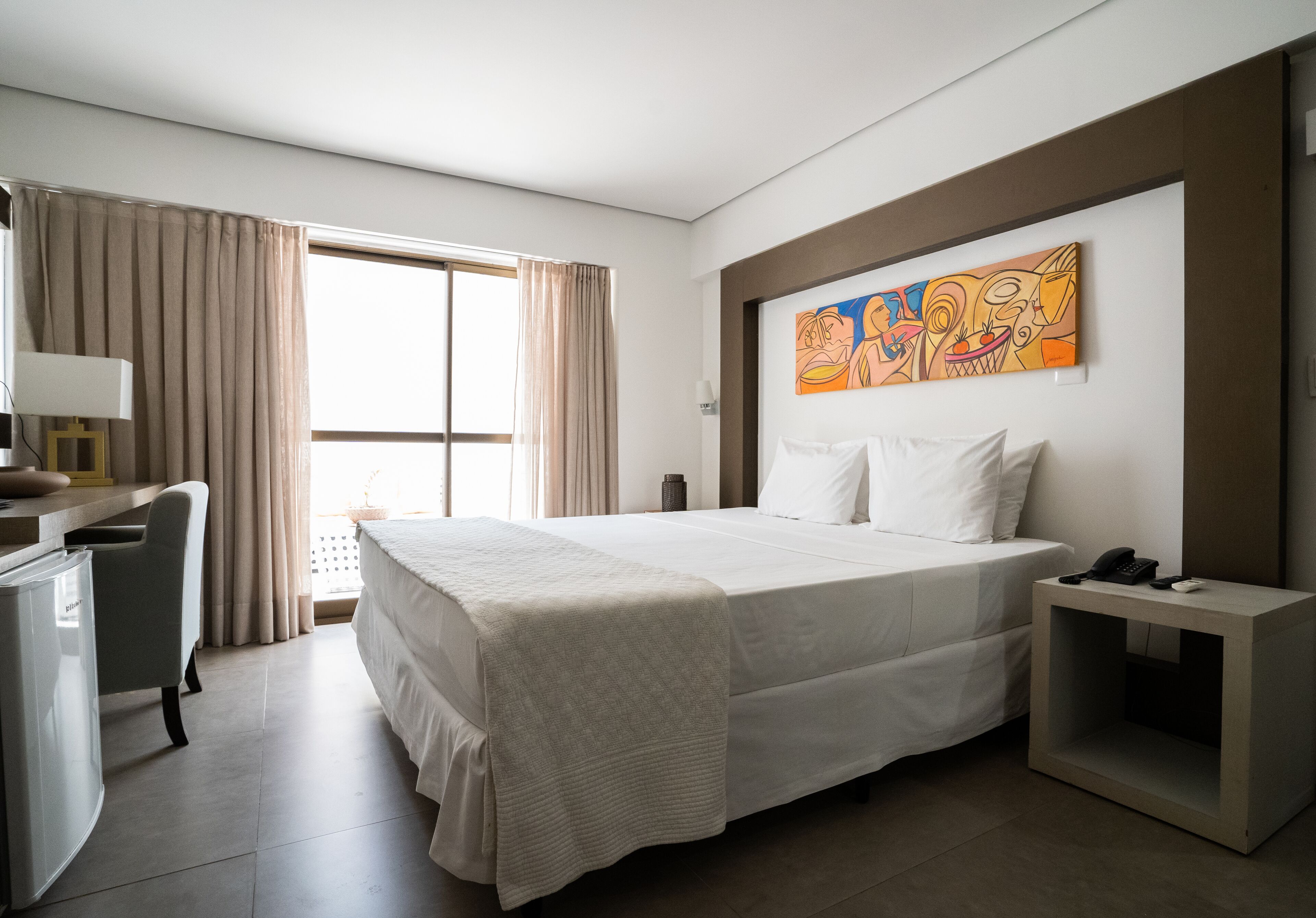 A hotel room with a large bed, a desk with a lamp, a small side table with a phone, and a window with curtains.