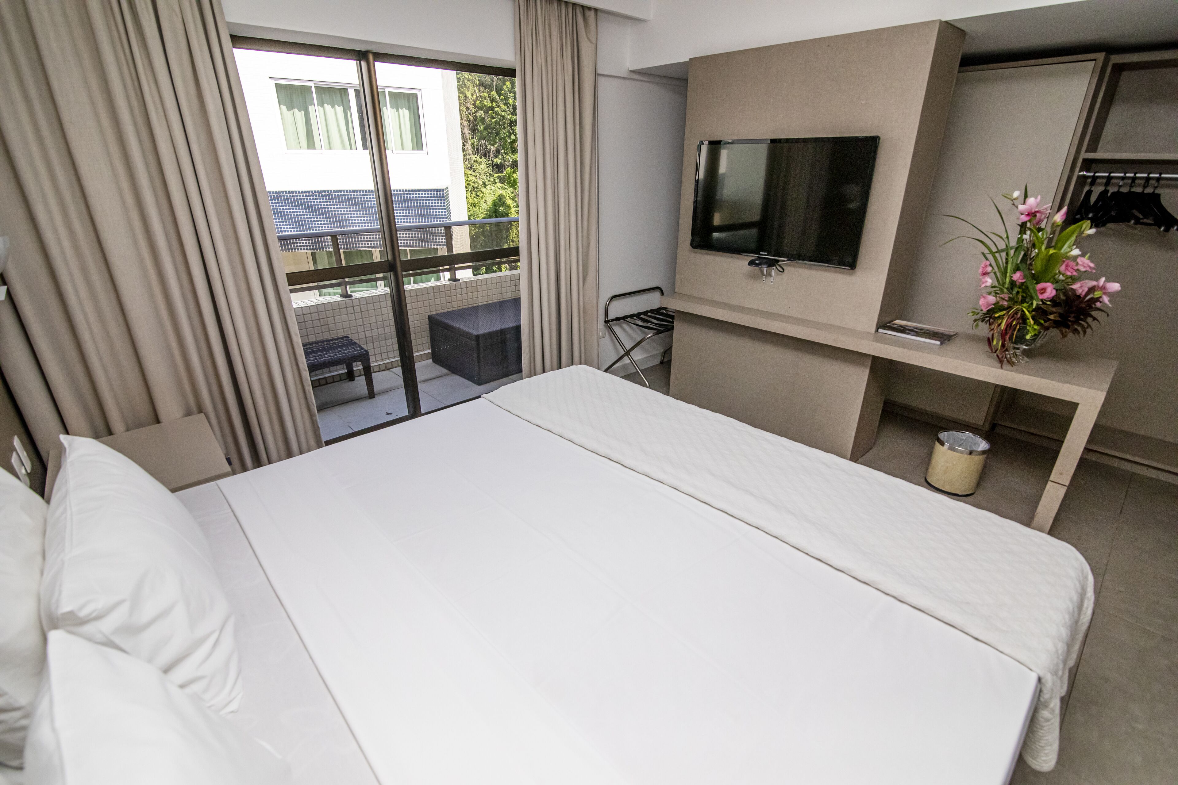 Une chambre d’hôtel avec un grand lit, un téléviseur fixé au mur, une petite table avec une chaise et un balcon donnant sur un paysage verdoyant.
