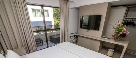 Une chambre d’hôtel avec un grand lit, un téléviseur fixé au mur, une petite table avec une chaise et un balcon donnant sur un paysage verdoyant.