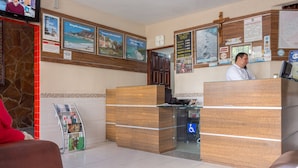 Ein Empfangsbereich mit einem Holzbüro, einer Uhr und einem Fernseher, der eine Person zeigt. An der Wand befinden sich gerahmte Bilder und ein Schild für Rollstuhlgerechter Zugang.