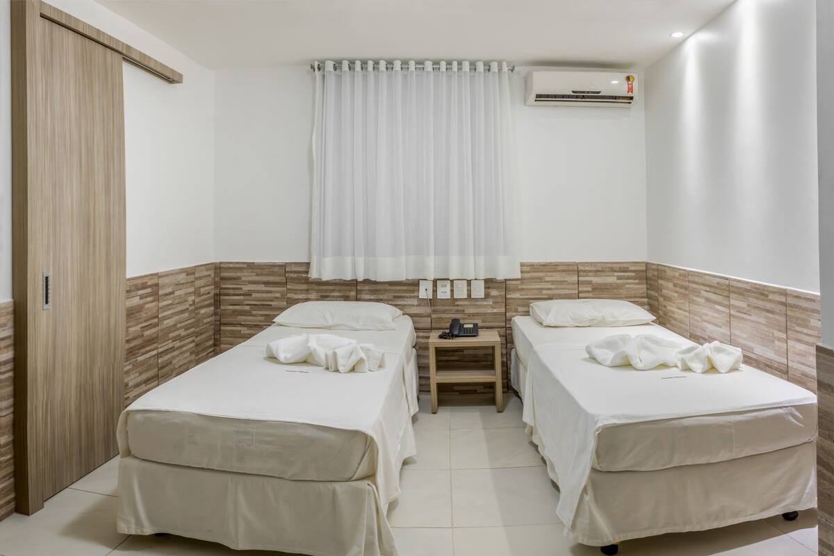 A hotel room with two beds, a wooden wardrobe, a nightstand with a phone, and an air conditioning unit.