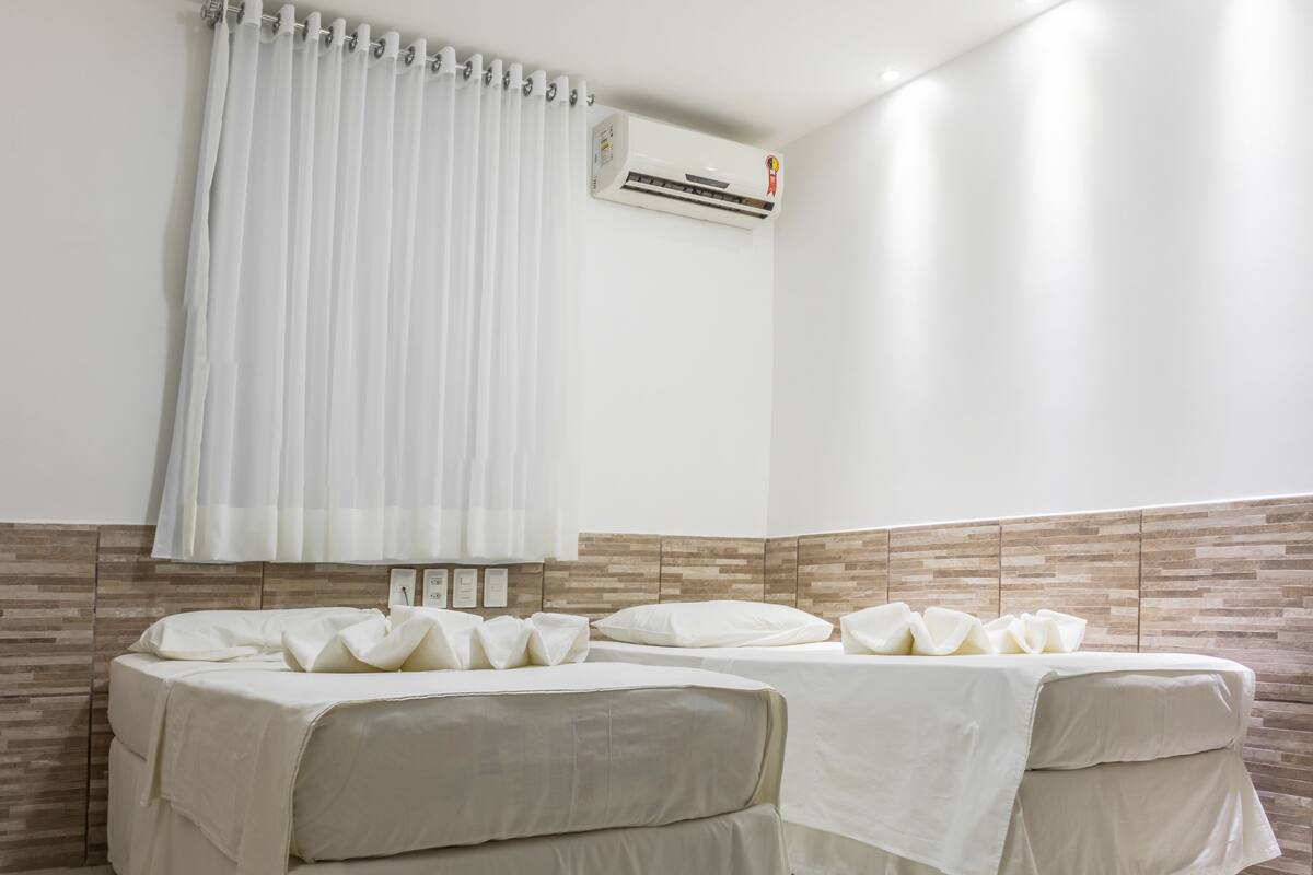 A hotel room with two beds, a white curtain, an air conditioning unit, and a tiled floor.