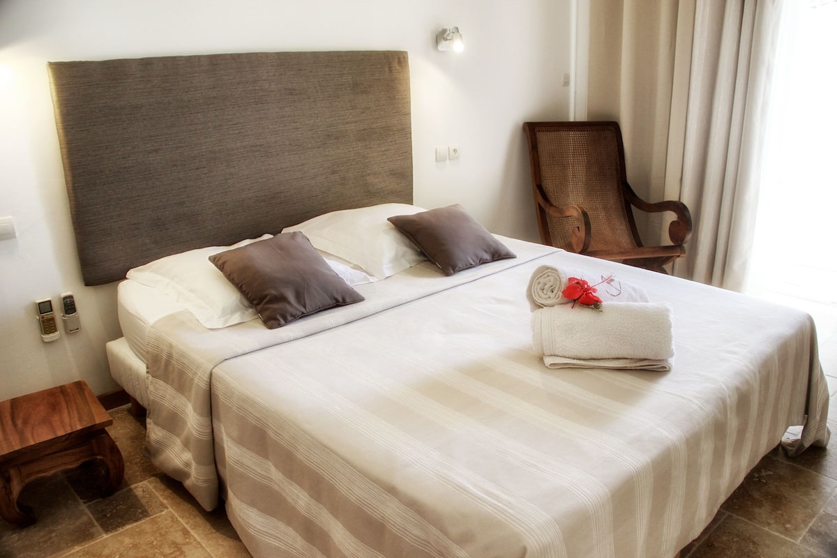 A neatly made bed with a striped bedspread, two pillows, and a rolled towel with a red flower on a hotel room.