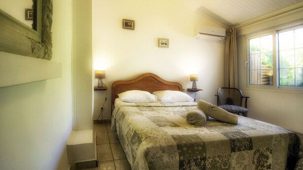 A neatly made bed with a patterned bedspread, two bedside lamps, a wooden headboard, and a chair by the window. - Hotel Villa Morgane (Saint-Pierre)