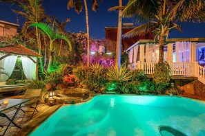 Un área de piscina nocturna con vista despejada a la piscina, la vegetación circundante y un pequeño pabellón.
