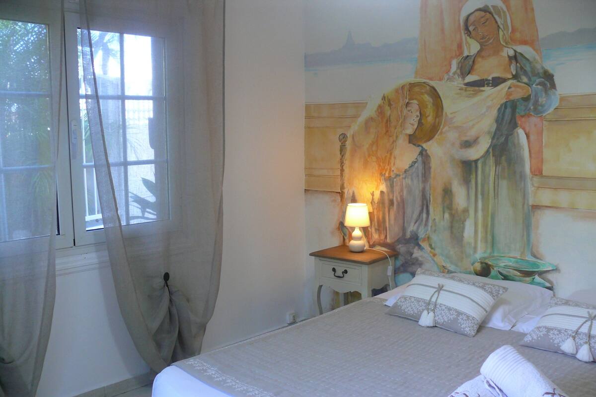 A hotel room with a bed, a nightstand with a lamp, a painting on the wall, and a window with curtains.