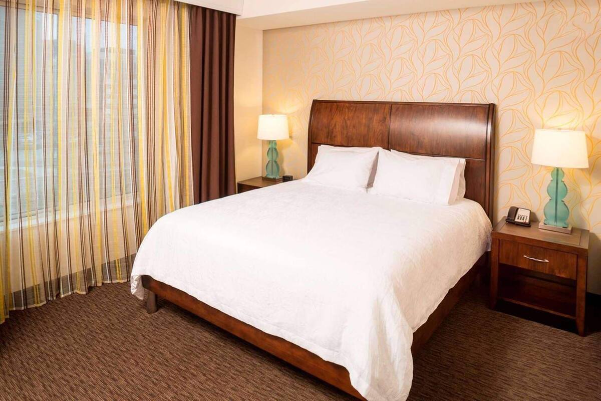 A hotel room with a large bed, two bedside tables with lamps, a window with curtains, and a patterned wall.
