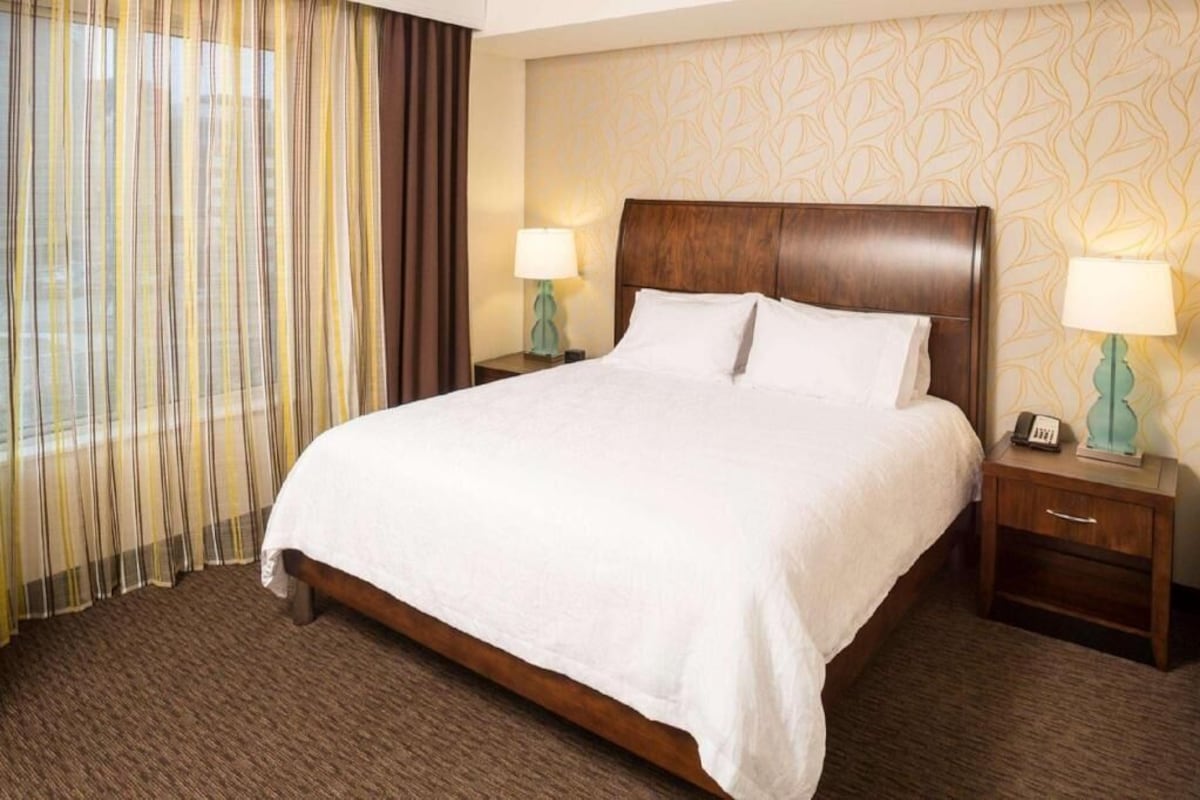 A hotel room with a large bed, two bedside tables with lamps, a window with curtains, and a patterned wall.