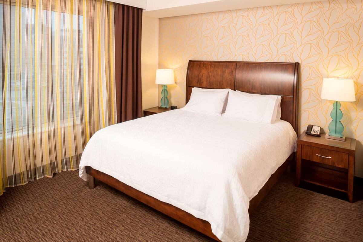 Une chambre d’hôtel avec un grand lit, une tête de lit en bois, deux tables de chevet avec des lampes, une fenêtre avec des rideaux et un papier peint à motifs.