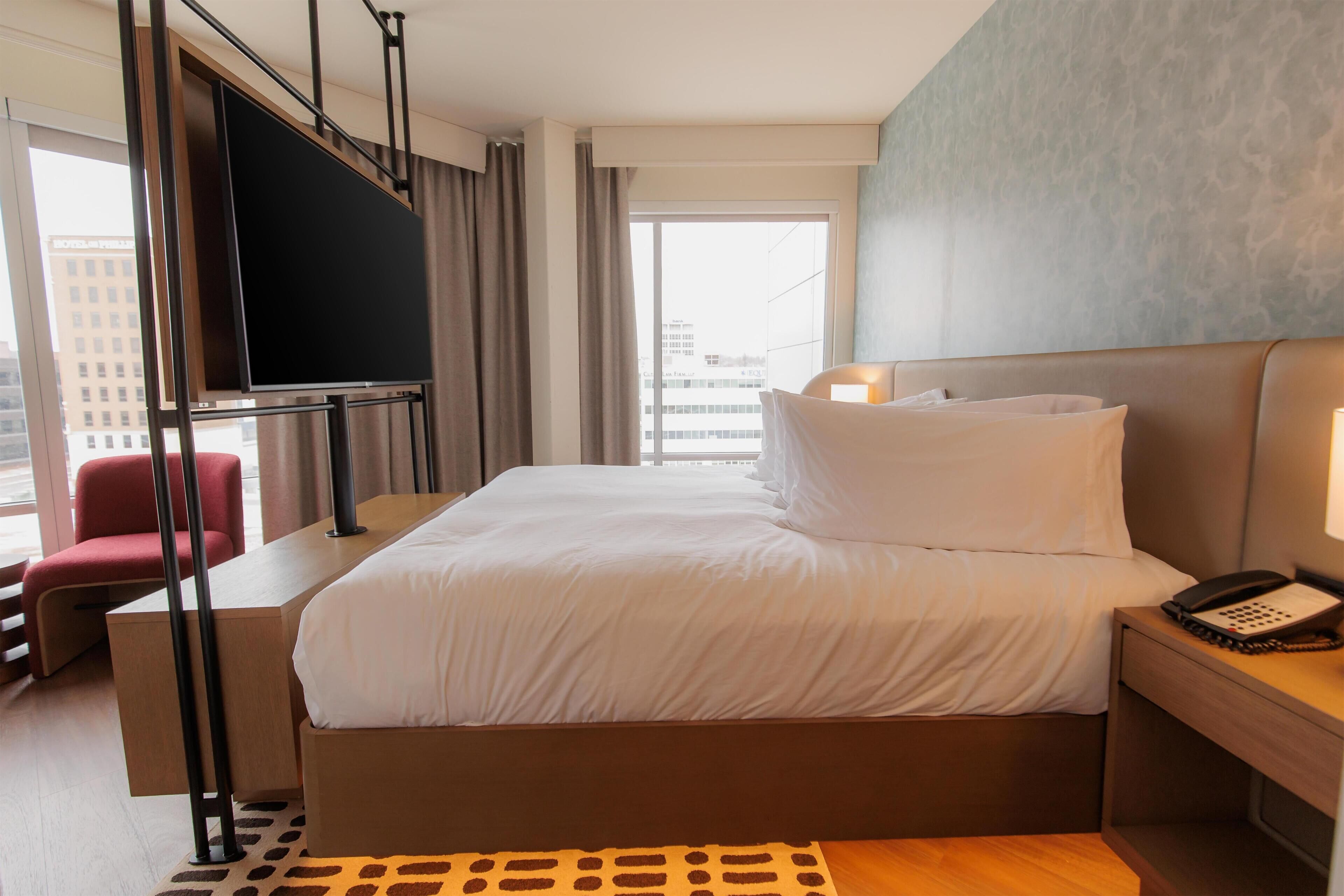 Habitación de hotel con cama, escritorio, silla, televisor, teléfono y ventana con vista a la ciudad.