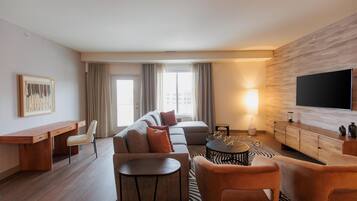 A modern hotel room with a sofa, armchairs, a desk, a flat-screen TV, and a patterned rug.