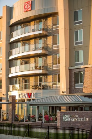 Un hotel de varios pisos con un diseño moderno, con balcones en cada planta y un restaurante visible llamado "CRAVE" a nivel de calle.